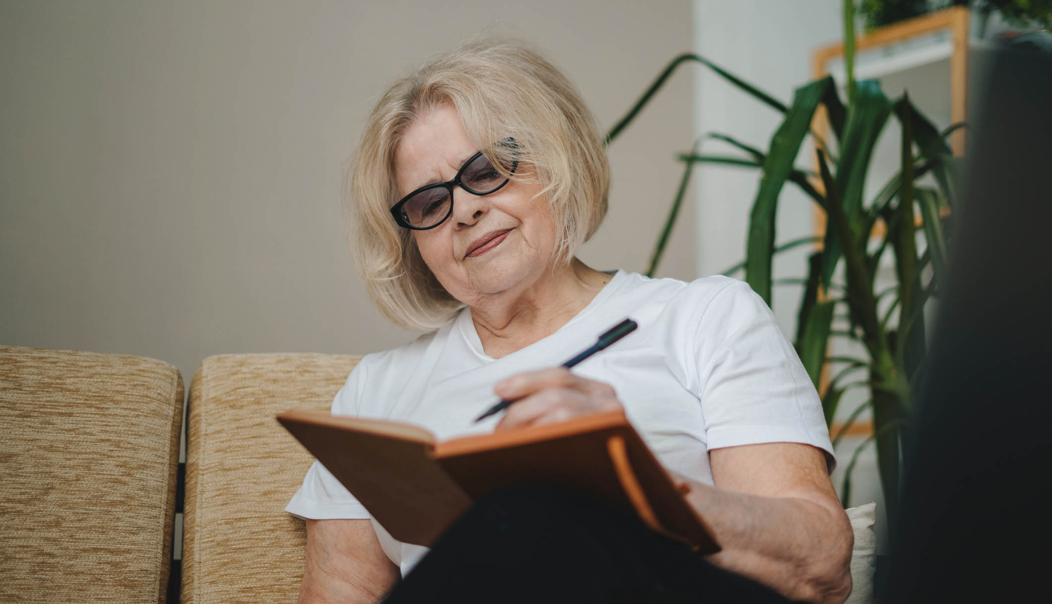 Happy senior woman with gray hair sitting on sofa swriting her life story